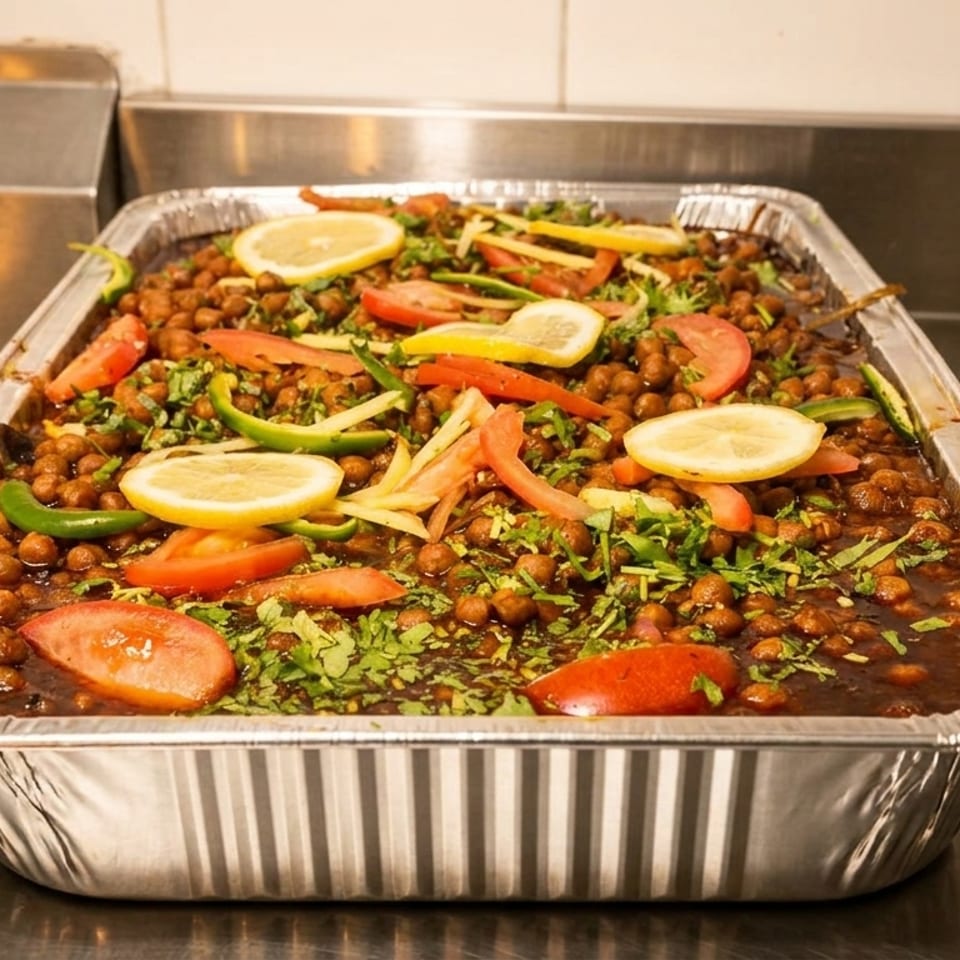 Aluminum catering tray filled with chickpeas in rich sauce, garnished with lemon slices, tomatoes, peppers, and cilantro.